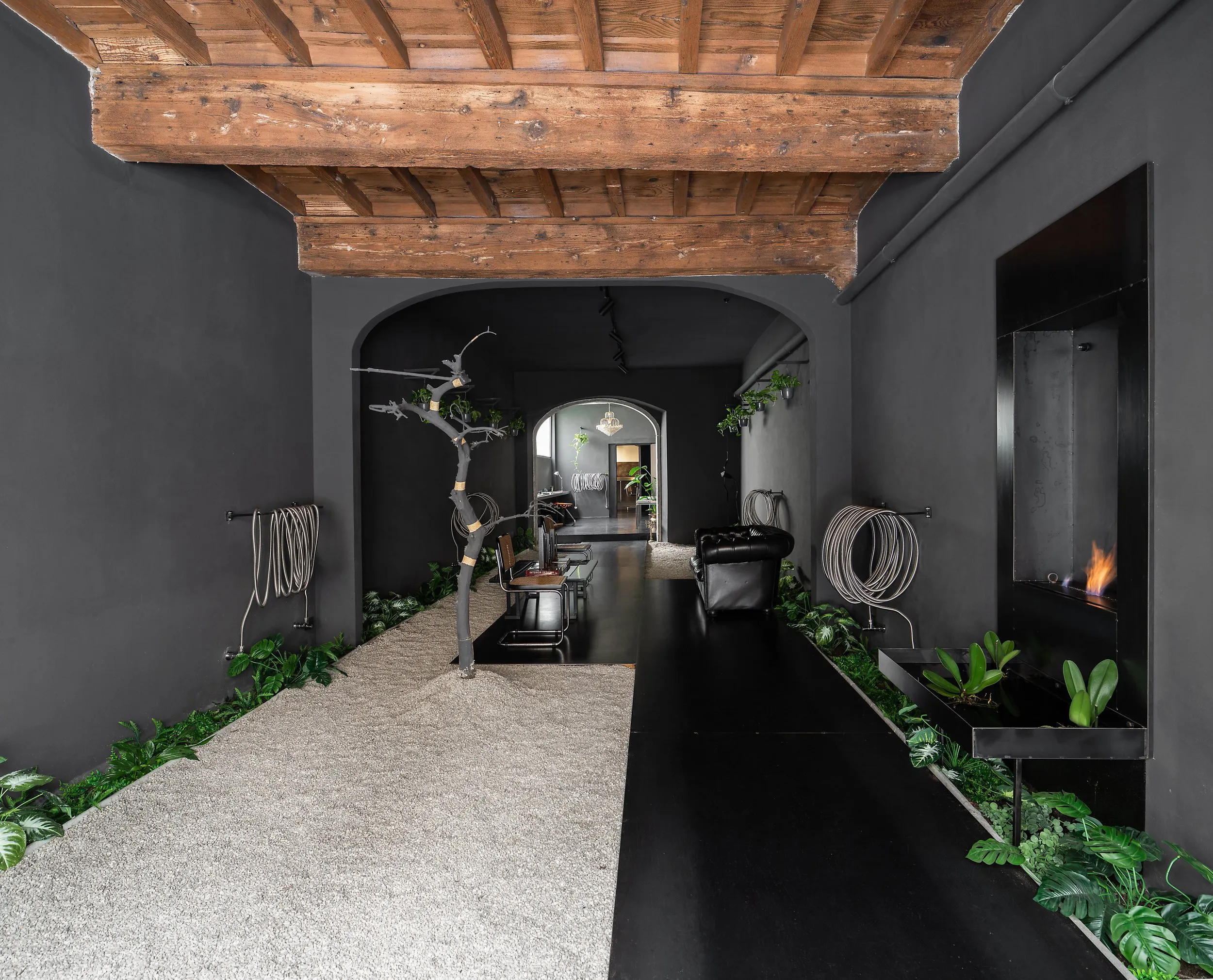 The atelier entrance: a gravel floor, the blackened olive trunk, the iron bio-fireplace alight and a Chesterfield sofa under the timber ceiling