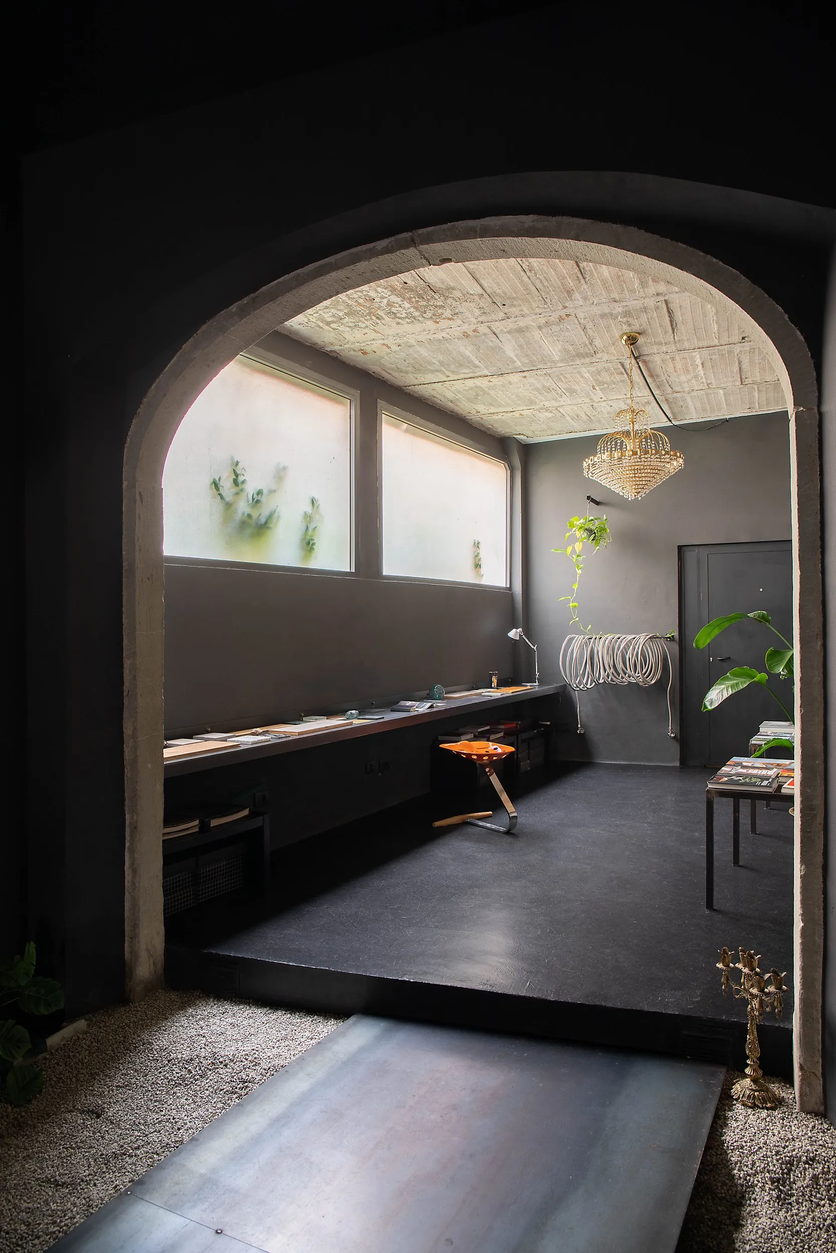 The materials library seen through the pietra serena arch: samples laid along a metal shelf, the orange Mezzadro stool, a crystal chandelier