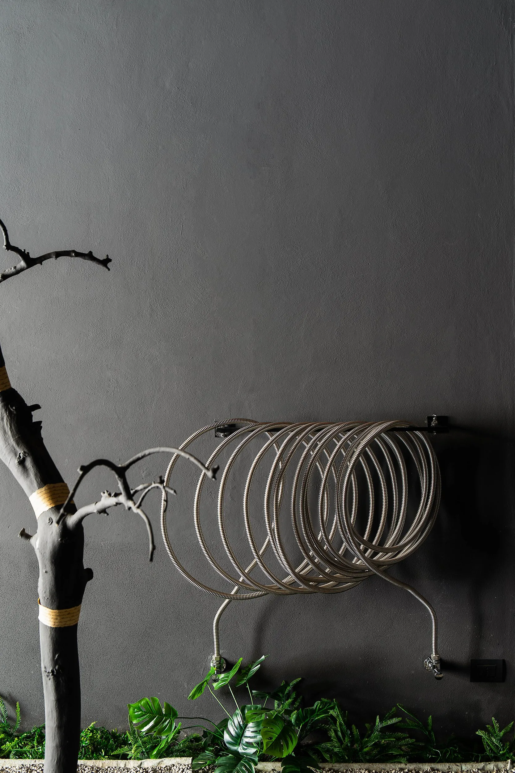 A radiator made of hydraulic pipes, coiled on the grey wall beside the blackened olive trunk bound in pale rope