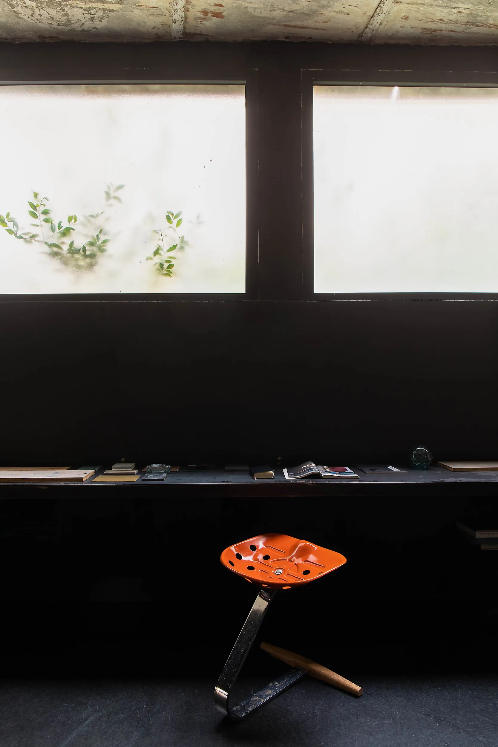 The desk: a single metal shelf running the length of the room under frosted windows, with the orange Mezzadro stool