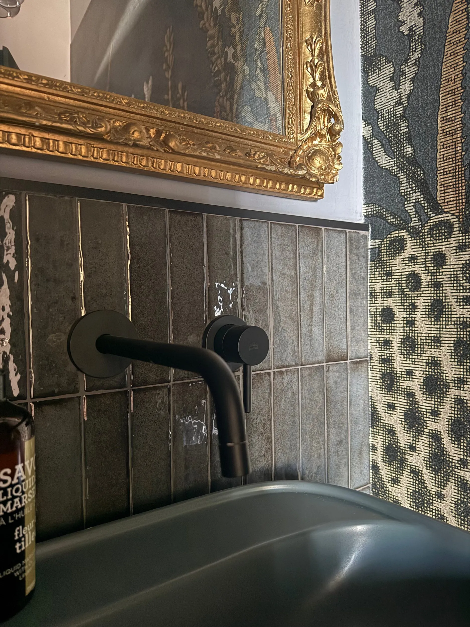 Black wall-mounted taps on dark glazed tiles, a gilt frame above