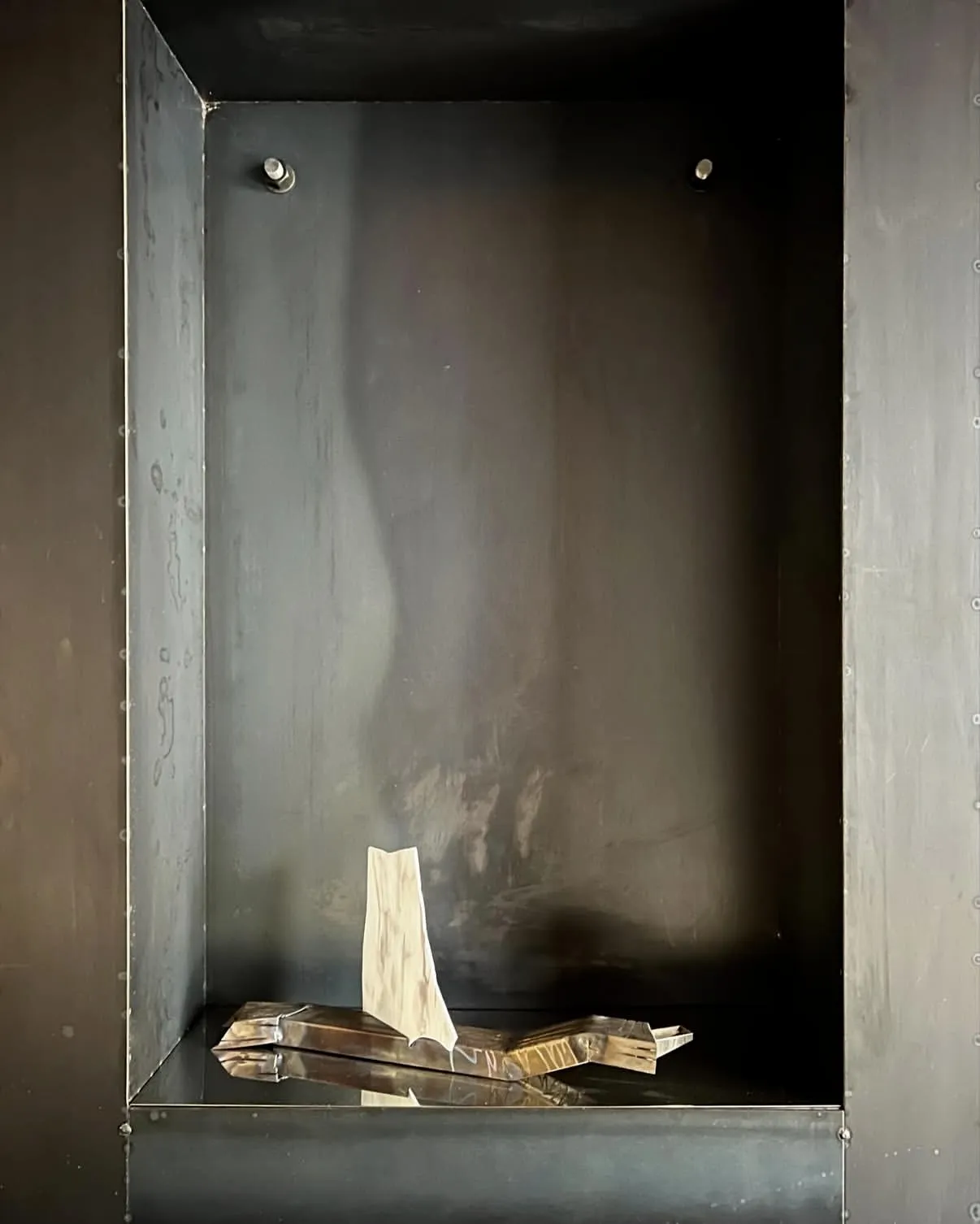 Inside the riveted iron niche: a pale sculpture standing on folded brass, alone in the dark box